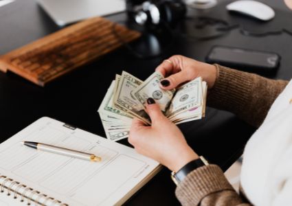 A Vienna landlord with black nail polish and a dark watch counts American money at a desk, above a pen and planner.